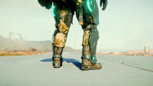 Shot from leg-level, from behind, someone's stood on a grey road that stretches out ahead. Desert and rocks in the distance. The person is wearing sandy coloured trousers and a metallic leg frame on the outside of their legs.
