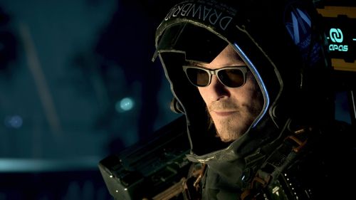 Close up of a man's face looking across to the left. He is wearing a large grey hood and black shades. It is dark but his face is lit up from a small source outside of shot.