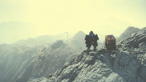 A man stands at a peak of a mountain range looking down towards a slim tower object in the distance on the next mountain peak. The sky is glaring. He wears a silver overall suit and carries behind him yellow cargo boxes on a floating carrier. 