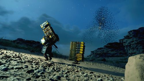 A man walks up a rocky hill at night. He is wearing a dark overall outfit. He carries a tall set of cargo boxes on his back and pulls behind him a floating carrier that is filled with similar yellow boxes of cargo. The surrounding area is rocky, suggesting a mountainous region. In the not-too-distant sky, a flock of black flying creatures similar to birds or bats circle together. 
