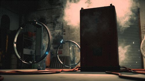 A smokey room in a factory. Metal roller shutter behind the large, metallic, rectangular machine. To the left side, two large circular metal rings with a single blue glowing light on each. 