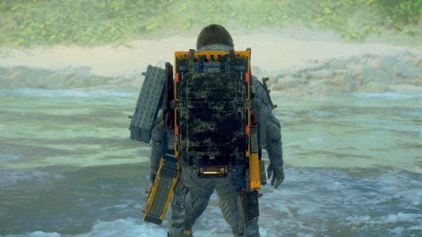 A man in a thick grey overall body suit and backpack with a yellow frame. He stands with his back to the camera, facing a river that passes in front of him.