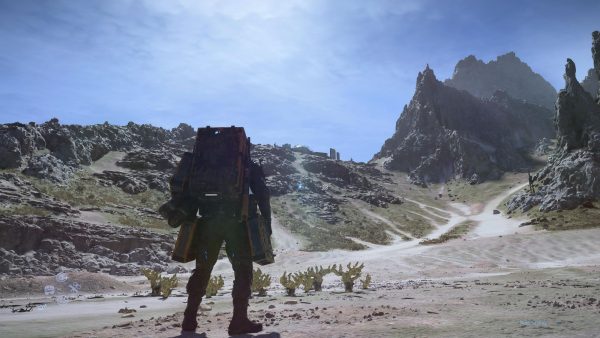 A man in a thick grey overall body suit and backpack with a yellow frame. He stands with his back to the camera, facing up a steep hill to the nearby summit. The sun's glare overhead in the almost-clear blue sky.