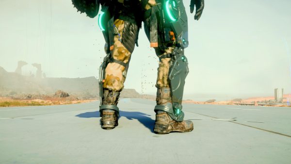 Shot from leg-level, from behind, someone's stood on a grey road that stretches out ahead. Desert and rocks in the distance. The person is wearing sandy coloured trousers and a metallic leg frame on the outside of their legs.