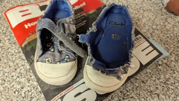 A pair of childs velcro-fastening shoes. Blue with white toes and blue cheater print pattern. One of the shoes has had its entire top section torn off by a very naughty dog