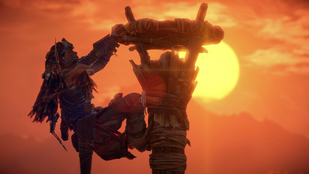 Top of a climbable post with the strong orange sun about the set in the background. The silhouette of a long-haired woman in make-shift armour, Aloy, hangs off the top.